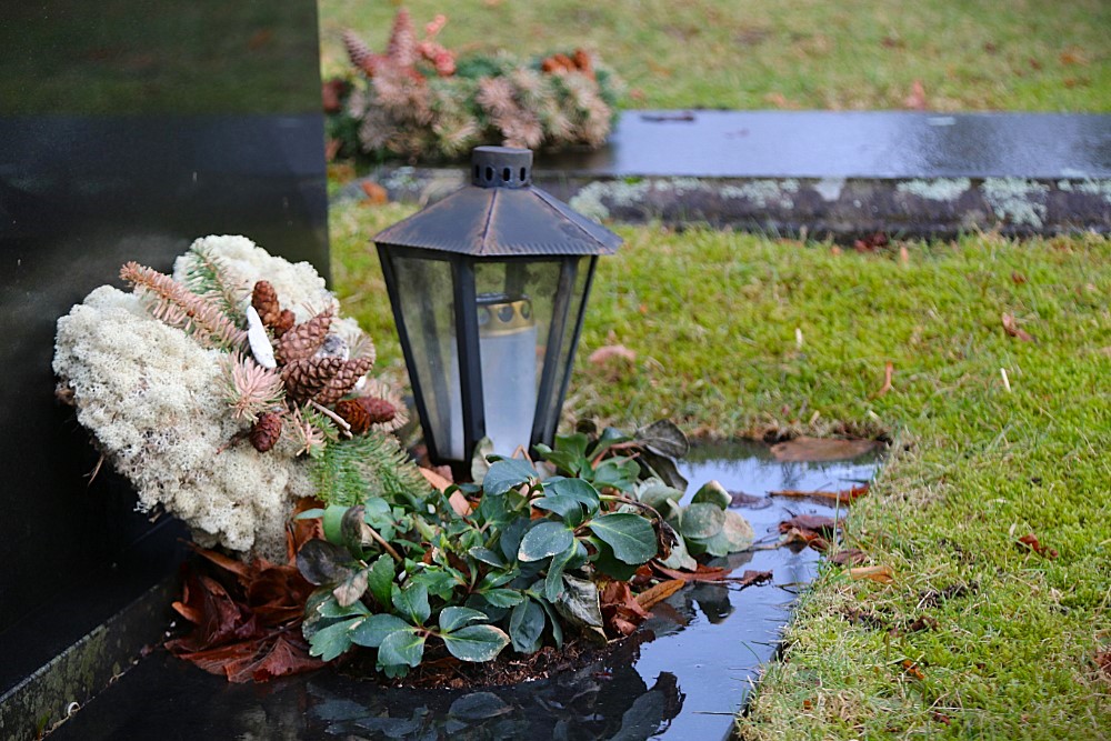 Gravsted med dekorasjon og lykt. En svart gravstein står til venstre, med en dekorasjon av hvit mose, kongler og granbar plassert ved foten. Ved siden av står en metalllykt, og foran er det grønne planter. Gress og våt jord omkranser området, og bak skimtes flere gravdekorasjoner. Foto